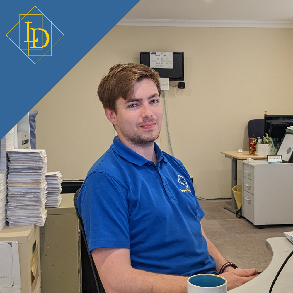 Aaron is a sitting at his desk with his hands in his lap smiling at the camera. Parts of the office can be seen in the background, some clutter is on his desk in the foreground. Up in the top left corner is the Logical Developments logo in gold on a blue background. The Logical Developments logo which is a diamond with the letters L and D inside of it. The letters are offset from each other vertically while also overlapping horizontally. The each sit inside of their own rectangle.