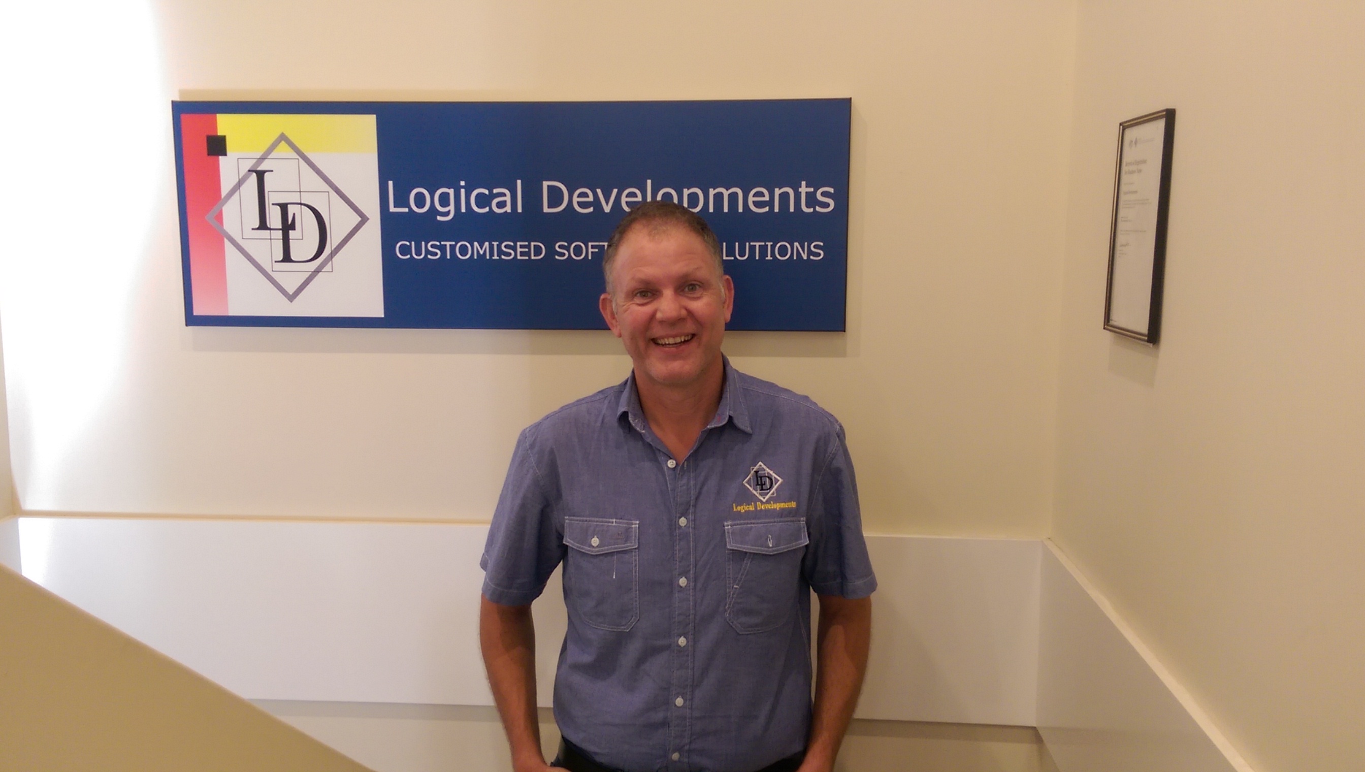 A picture of Shane. He stands beneath the Logical Developments banner that is hanging up in the stair case up the office. He wears the dress shirt that is part of the uniform and is smiling.